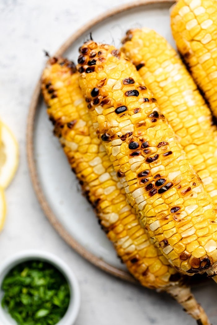 Plate with a stack of grilled corn on the cob on it. Next to it is a small bowl of herbs.