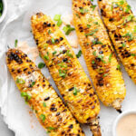Grilled corn on a plate topped with sauce and green onions. Next to it a small bowl of more sauce and a small bowl of green onions.