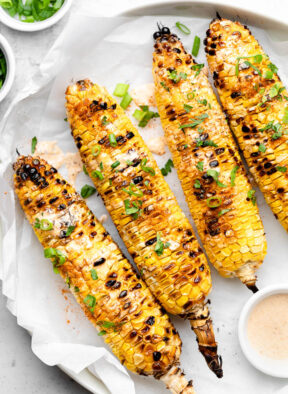 Grilled corn on a plate topped with sauce and green onions. Next to it a small bowl of more sauce and a small bowl of green onions.
