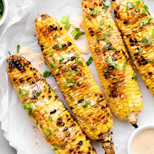 Grilled corn on a plate topped with sauce and green onions. Next to it a small bowl of more sauce and a small bowl of green onions.