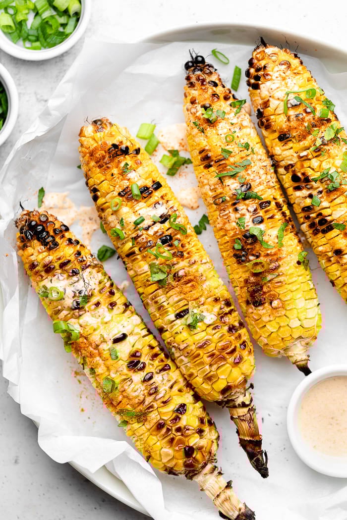Grilled corn on a plate topped with sauce and green onions. Next to it a small bowl of more sauce and a small bowl of green onions.