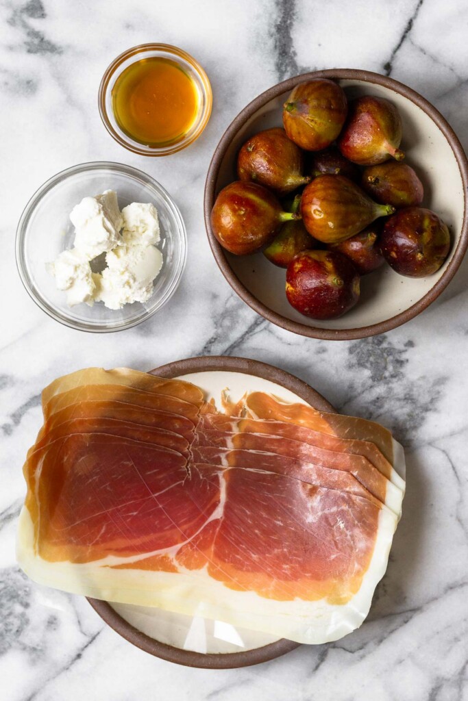 White marble counter with a bowl of fresh figs, a plate of slices of prosciutto, a bowl of goat cheese, and a bowl of honey,