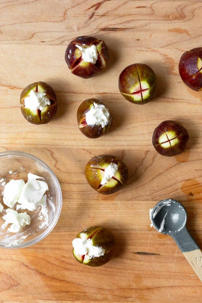 Wooden cutting board with fresh figs with cross slices across the top of them. Some are stuffed with goat cheese. There is a bowl of goat cheese and a teaspoon on the cutting board.