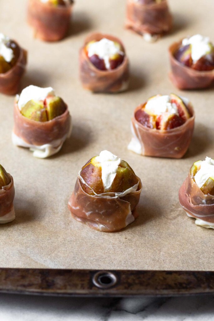 Fresh figs stuffed with goat cheese and wrapped in prosciutto on a baking sheet lined with parchment paper.