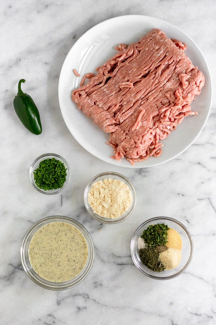 White counter with a plate of raw ground turkey. bowl of spices. bowl of coconut flour, bowl of ranch, bowl of fresh chopped chives, and a jalapeño.