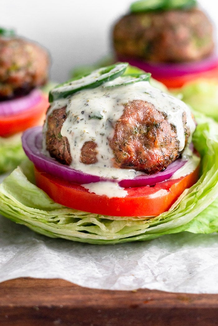 Jalapeño ranch turkey burger with ranch topped with sliced jalapeños and ranch dressing. It is on a lettuce cup with sliced tomato and red onion with more burgers behind it.
