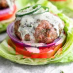 Ranch turkey burger covered in ranch dressing and topped with sliced jalapeños. It is in a lettuce wrap with tomato and red onion. Behind it is more burgers.