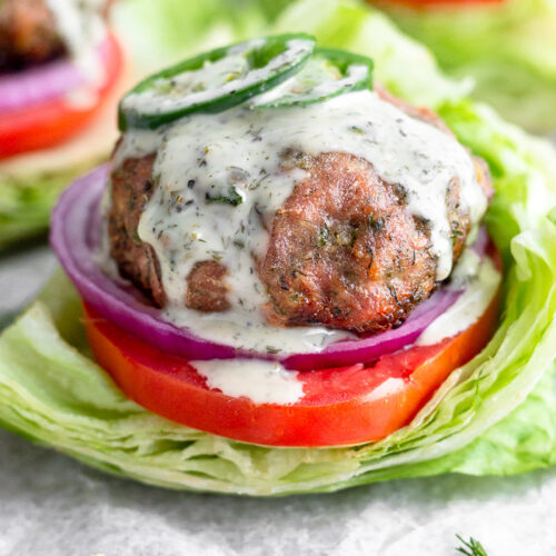 Ranch turkey burger covered in ranch dressing and topped with sliced jalapeños. It is in a lettuce wrap with tomato and red onion. Behind it is more burgers.