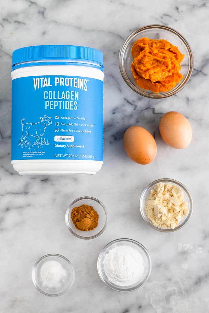 White counter with a blue container of collagen, bowl of pumpkin puree, 2 eggs, bowl of coconut flour, bowl of tapioca flour, bowl of baking soda, and bowl of spices.