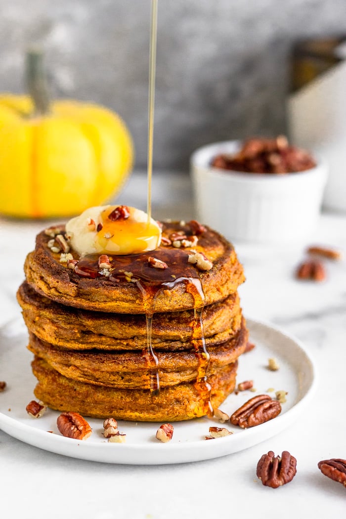 Stack of gluten free pumpkin protein pancakes with maples syrup being poured on them. Behind them is a small container of nuts, a mug, and a yellow pumpkin.