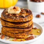 Stack of Protein Powder Pumpkin Pancakes on a white plate with maple syrup, coconut butter, and chopped nuts on top of them. Behind it is a mug, dish of nuts, and a yellow pumpkin.
