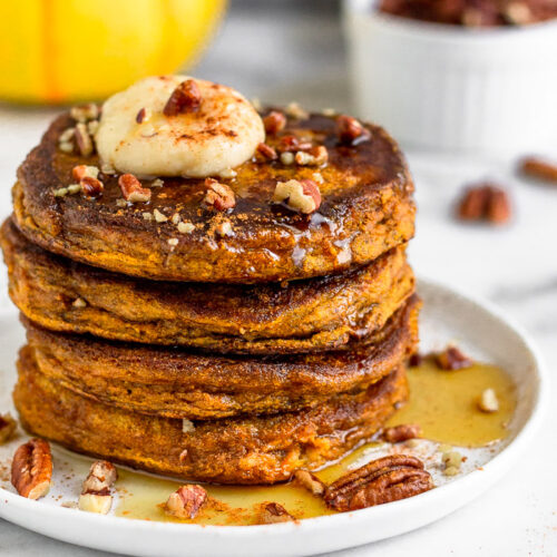 Stack of Protein Powder Pumpkin Pancakes on a white plate with maple syrup, coconut butter, and chopped nuts on top of them. Behind it is a mug, dish of nuts, and a yellow pumpkin.