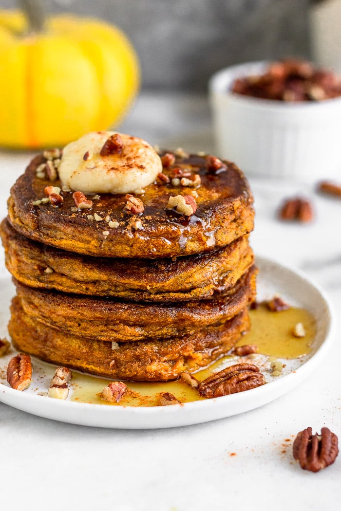 Stack of Protein Powder Pumpkin Pancakes on a white plate with maple syrup, coconut butter, and chopped nuts on top of them. Behind it is a mug, dish of nuts, and a yellow pumpkin.