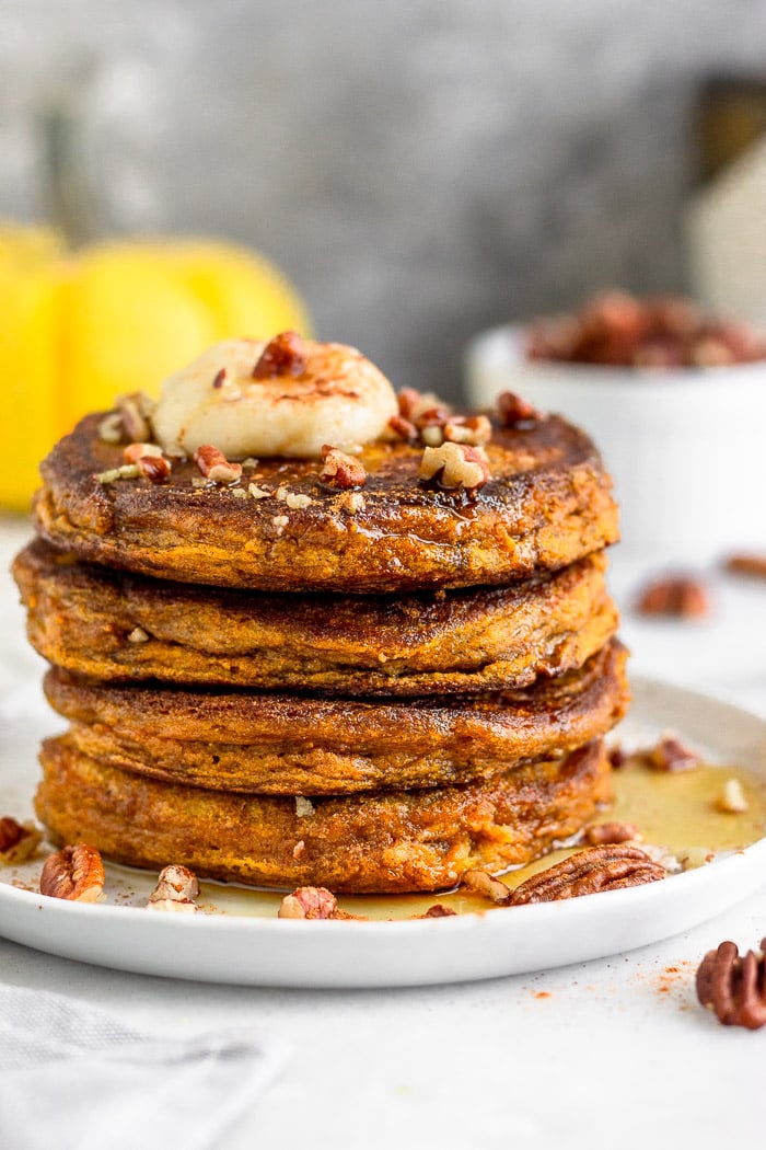 Stack of gluten free pumpkin protein pancakes sitting on a white plate. The pancakes are topped with coconut butter and chopped nuts and there is syrup all over the plate. Behind it is a yellow pumpkin and a small dish of nuts.