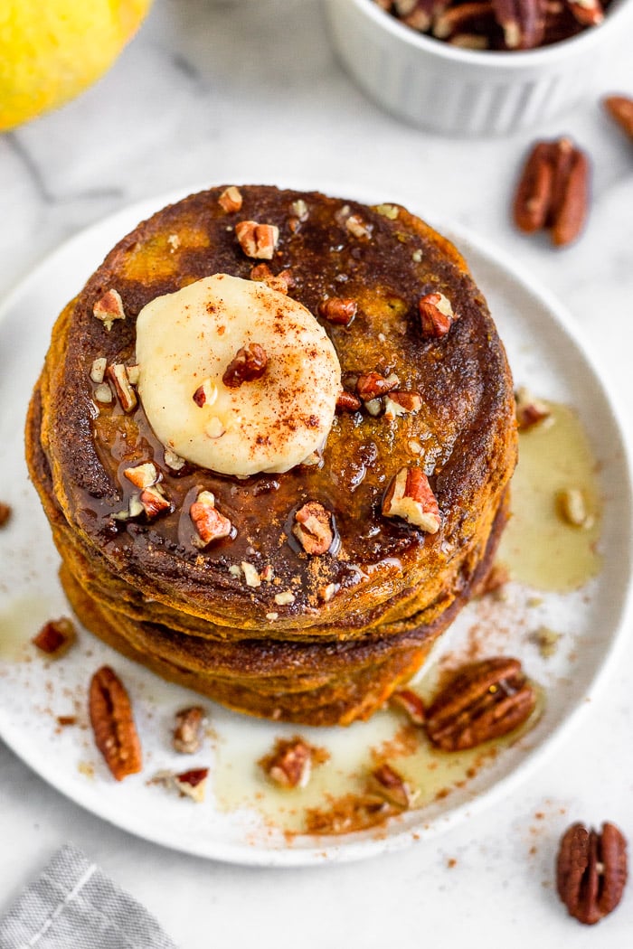 Overhead shoot of a stack of healthy pumpkin protein pancakes topped with coconut butter, chopped pecans, and maple syrup. Next to it is a bowl of pecans with more pecans next to it.