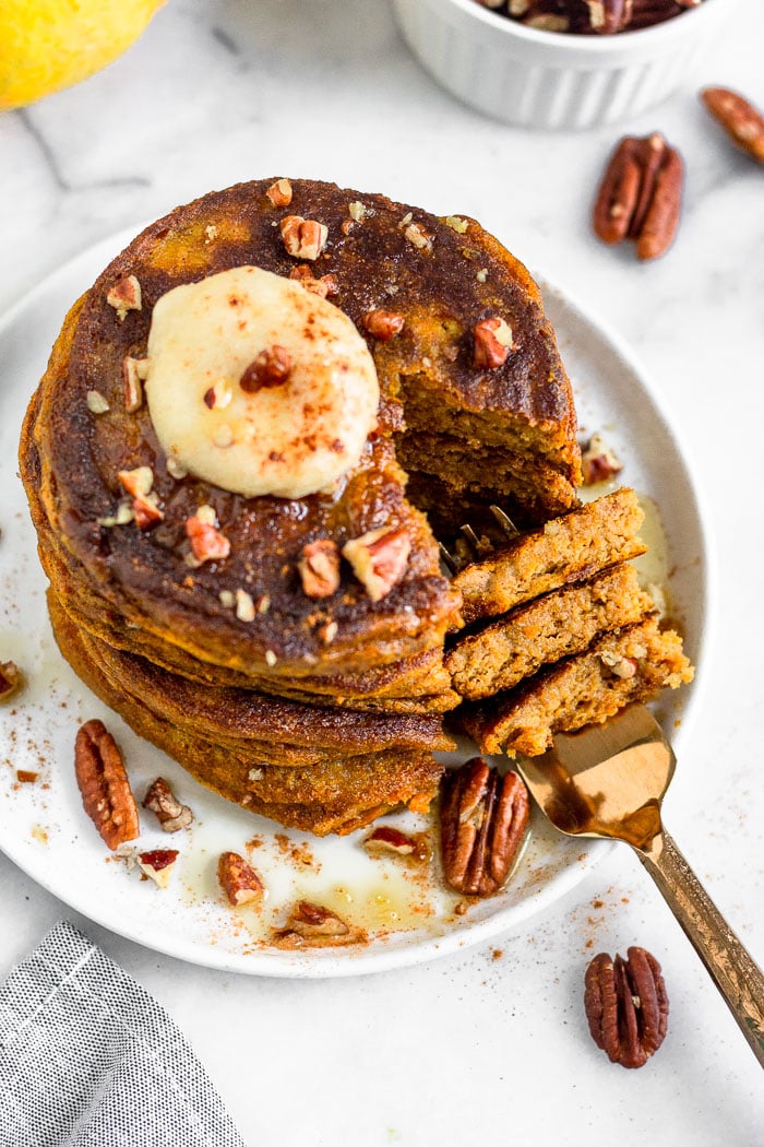 Overhead view of a stack of pumpkin pancakes with protein topped with coconut butter, chopped pecans, and maple syrup. A slices is missing from the stack and is on a fork that is laying on the plate.