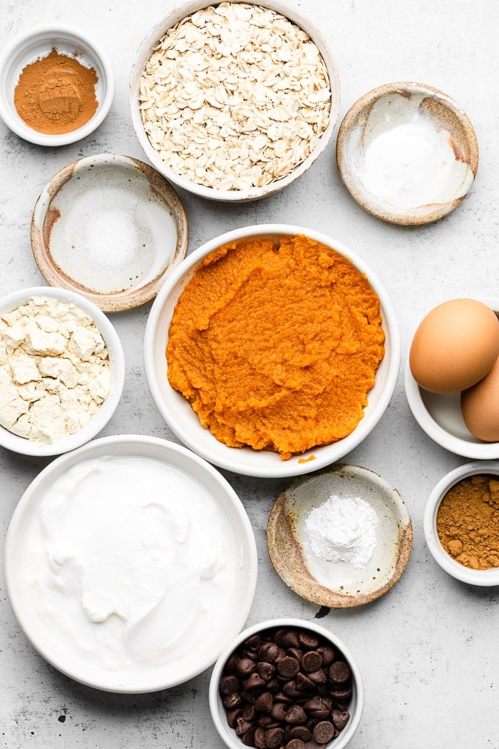 White counter filled with a bowl of spices, bowl of rolled oats, bowl of baking soda and powder, bowl of eggs, bowl of pumpkin puree, bowl of greek yogurt, and a bowl of protein powder.
