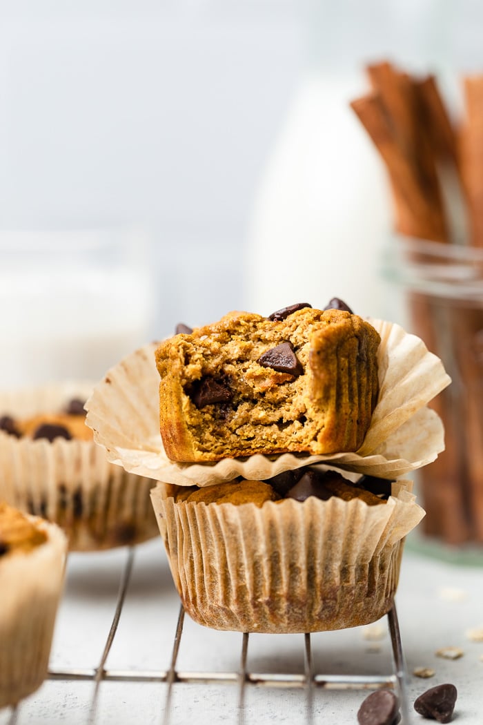 Two healthy pumpkin protein muffins stacked on top of each other with the top one with a bite taken out of it. Behind it more muffins, glass of milk, and cinnamon sticks. They are sitting on a cooling rack.