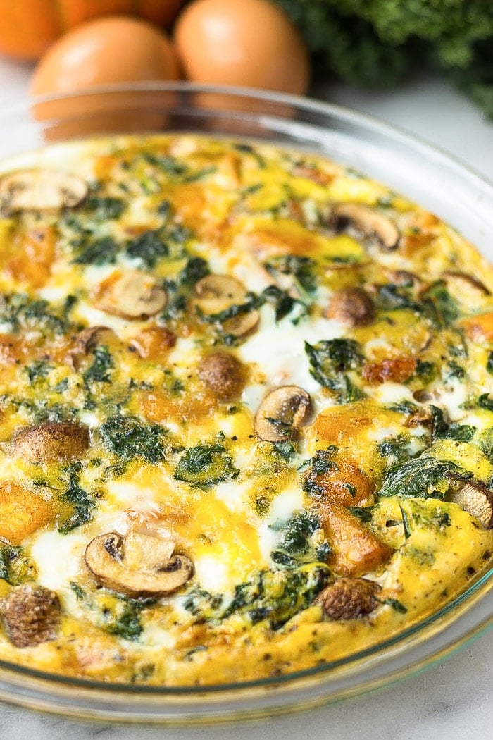 Crustless pumpkin quiche in a pie pan with eggs, pumpkin, and kale behind it.