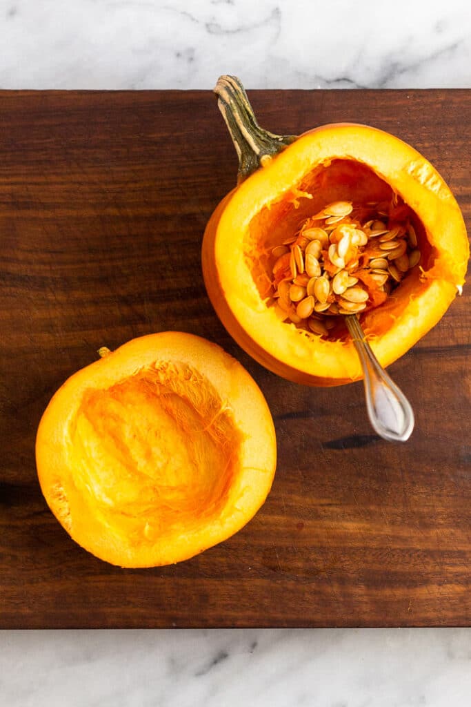 A small sugar pumpkin cut in half sitting on a cutting board. The is a spoon in one of the halves.