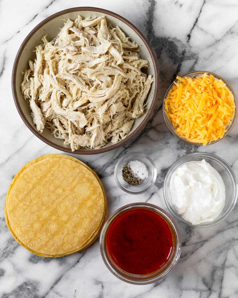 White marble counter with a bowl of shredded chicken, a bowl of shredded cheese, a bowl of greek yogurt, a bowl of buffalo sauce, a stack of corn tortillas, and a small bowl of salt and pepper.