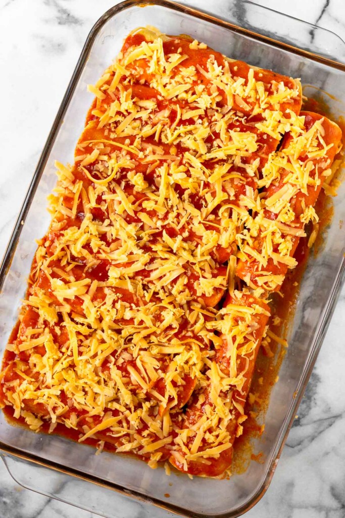 A glass baking dish of enchiladas topped with sauce and sprinkled with shredded cheese before it is baked.