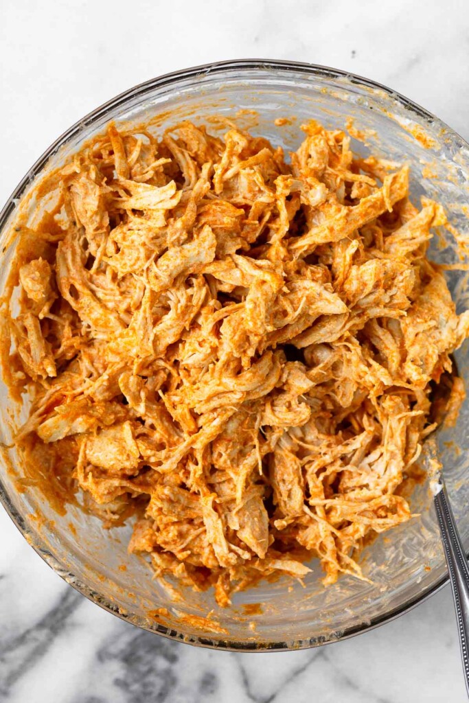 A bowl with shredded buffalo chicken in it. A spoon is sticking out of the bowl.