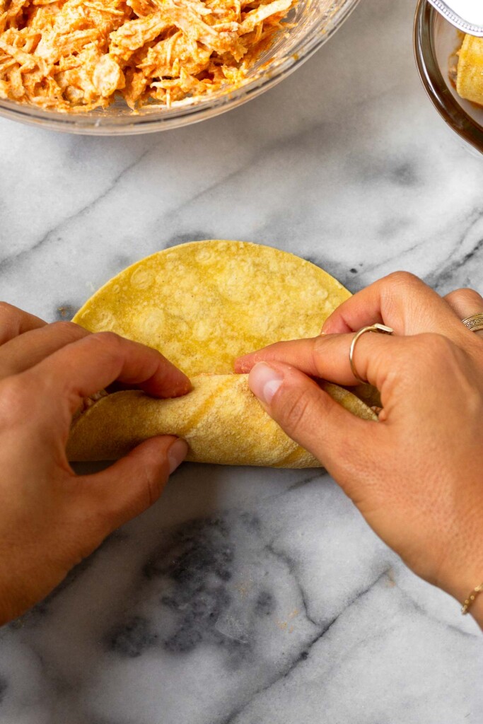 Two hands rolling up a chicken enchilada. Next to it is a bowl of shredded chicken.