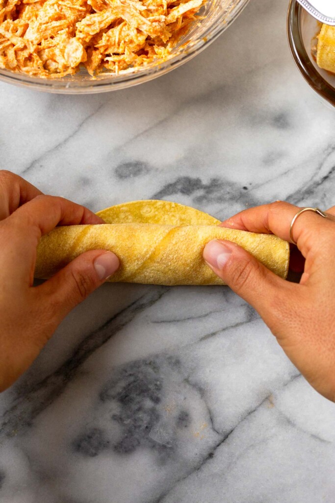 Chicken enchilada being rolled up. Next to it is a bowl of shredded chicken.