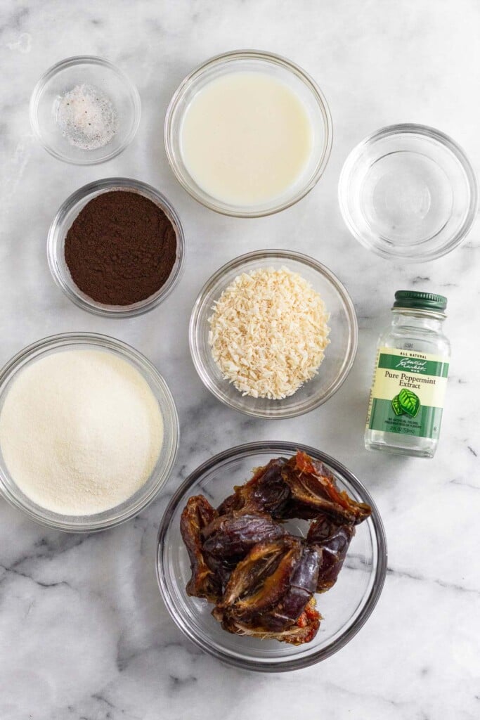 White marble counter with a bowl of coconut butter, a bowl of water, a small jar of peppermint extract, a bowl of shredded coconut, a bowl of medjool dates, a bowl of collagen, a bowl of cocoa powder, and a small bowl of salt.