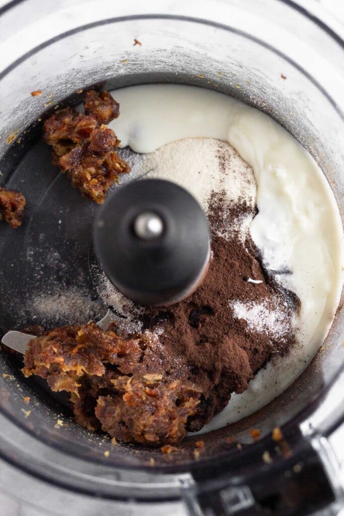 Food processor with broken up dates, cocoa powder, collagen, coconut butter, and salt in it.
