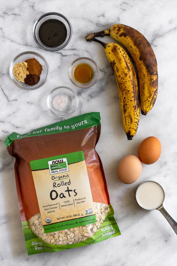 Overhead shot of two brown bananas, 2 brown eggs, small measuring cup of almond milk, bag of rolled oats, a couple bowls spices, small bowl of vanilla extract, and a small bowl of molasses.