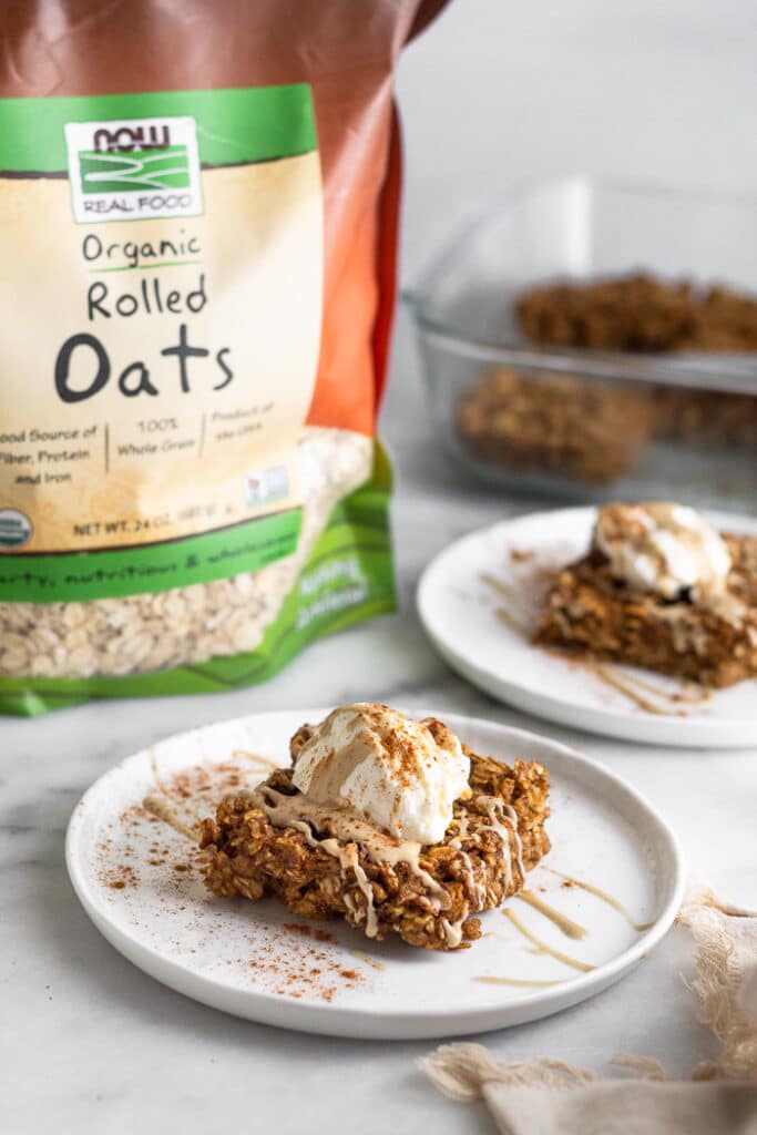 Two plates with baked gingerbread oatmeal bars on it. They are topped with yogurt, nut butter, and cinnamon. Next to them is a bag of rolled oats and large dish of more baked oatmeal.
