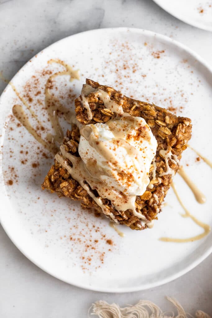 Overhead shot of a baked oatmeal bar on a plate topped with yogurt, nut butter, and cinnamon.