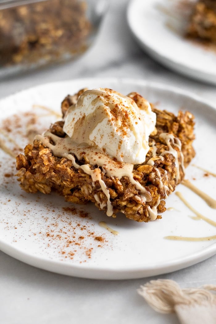 Gingerbread baked oatmeal bar on a plate topped with yogurt, nut butter, and cinnamon. Behind it is another plate and a pan of more baked oatmeal
