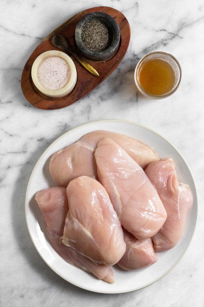 Marble counter top with two marble bowls with salta and pepper in them, a glass jar of broth, and plate of raw chicken breasts.