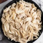 A black bowl filled with shredded chicken sprinkled with pepper. The bowl is on a striped kitchen towel with a wooden dish next to it.