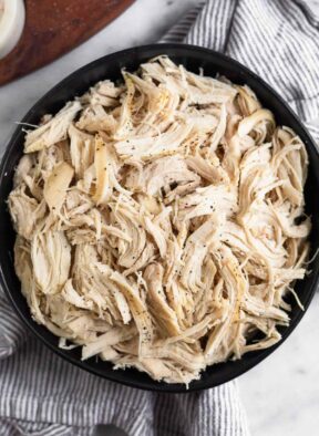 A black bowl filled with shredded chicken sprinkled with pepper. The bowl is on a striped kitchen towel with a wooden dish next to it.