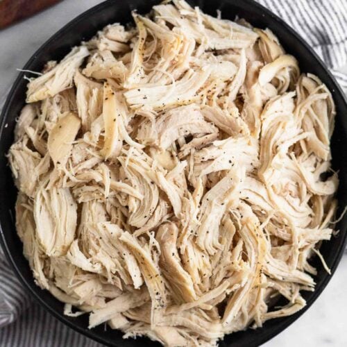 A black bowl filled with shredded chicken sprinkled with pepper. The bowl is on a striped kitchen towel with a wooden dish next to it.