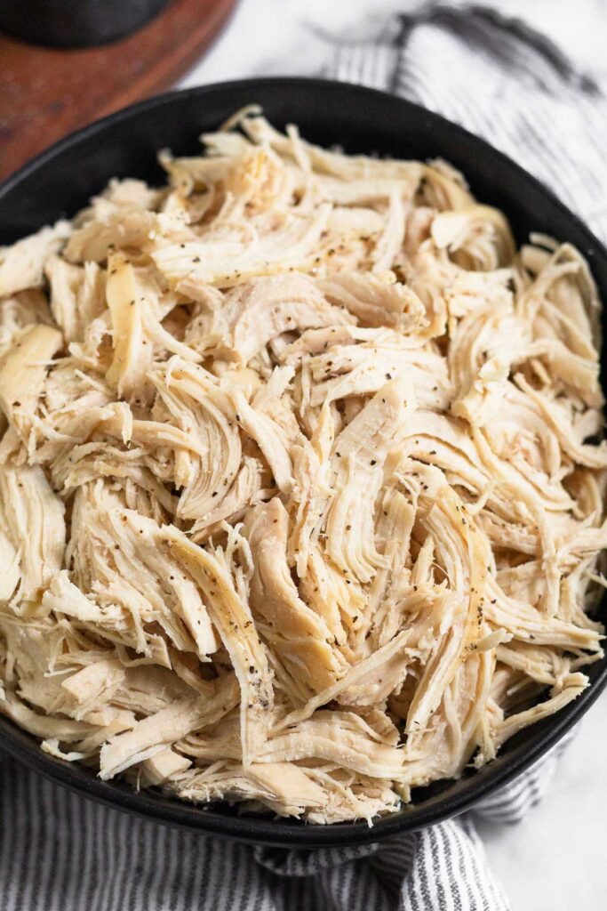 A close-up of a black bowl filled with shredded chicken sprinkled with pepper. The bowl is on a striped kitchen towel with a wooden dish in the background.