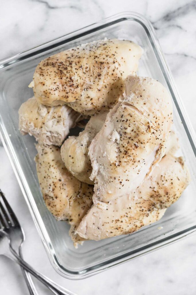 Rectangular glass container filled with cooked chicken breasts seasoned with pepper on a marble counter. Next to the dish is two forks.