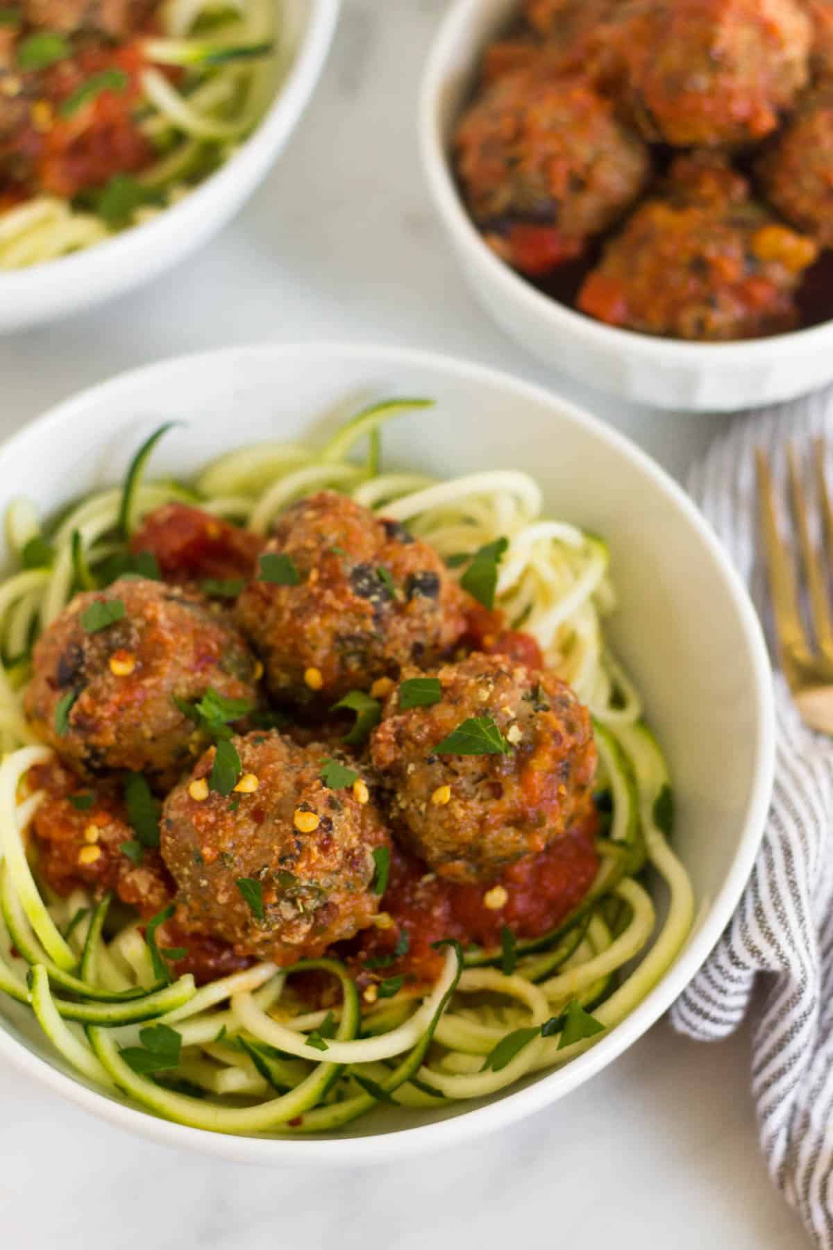 Bowl of zucchini noodles and supreme pizza meatballs with a smaller bowl of meatballs and sauce and another bowl of zucchini noodles in the background