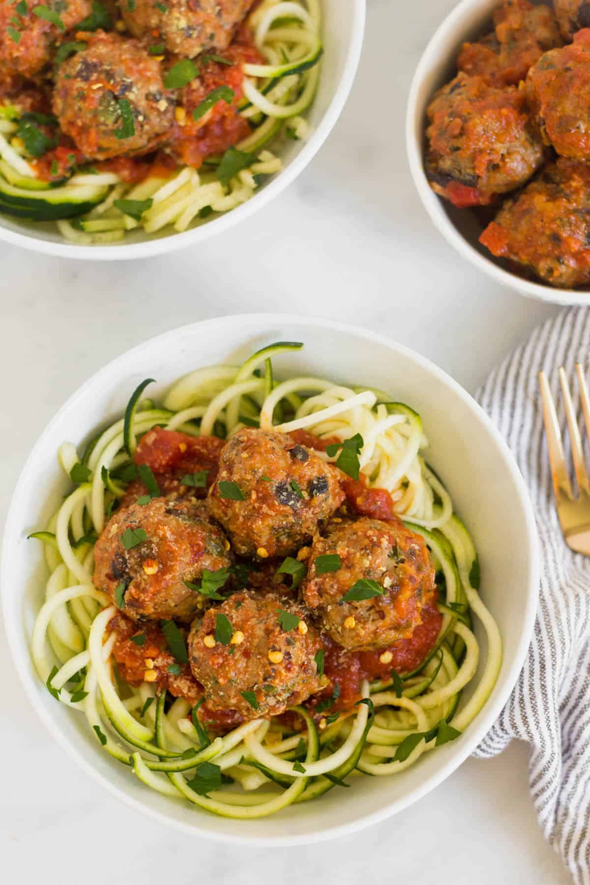 Overhead shot of two white bowls with zucchini noodles with meatballs and sauce and a small white bowl of meatballs and sauce