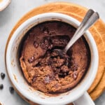 Overhead shot of a protein mug cake with a spoon in it with some of the middle scooped out. The mug is sitting on a small wooden coaster and around it are some mini chocolate chips.