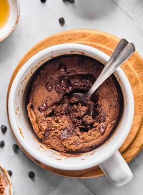 Overhead shot of a protein mug cake with a spoon in it with some of the middle scooped out. The mug is sitting on a small wooden coaster and around it are some mini chocolate chips.