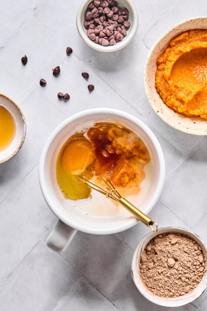 Overhead shot of a white cup with milk, pumpkin puree, an egg, maple syrup, and vanilla bean paste before it is mixed together. There is a small gold whisk in the mug too. Around the mug is a bowl of pumpkin puree, a bowl of cocoa powder, a bowl of maple syrup, and a bowl of mini chocolate chips with some around the bowl.