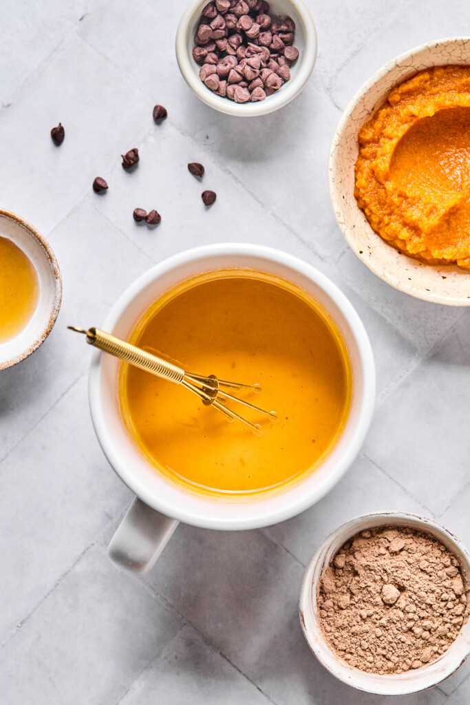 Overhead shot of a white cup with a wet batter and a small gold whisk in it. Around the mug is a bowl of pumpkin puree, a bowl of cocoa powder, a bowl of maple syrup, and a bowl of mini chocolate chips with some around the bowl.