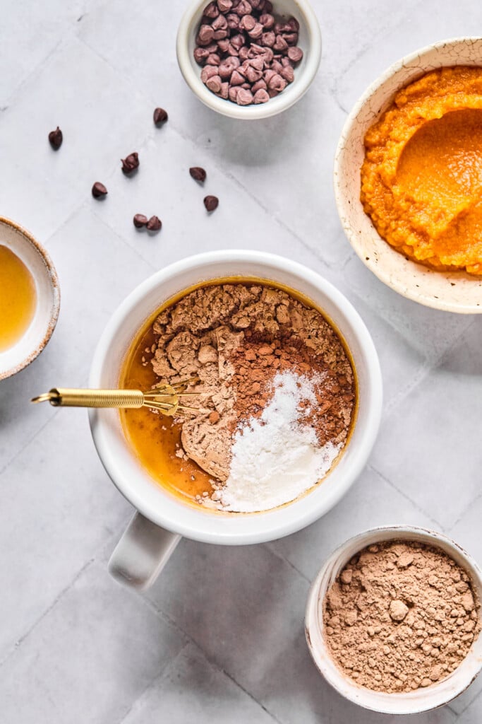 Overhead shot of a white mug with a wet batter with some chocolate protein powder, cocoa powder, salt, and baking powder overtop if it before it is mixed together. A small gold whisk is in the mug too. Around the mug is a bowl of pumpkin puree, a bowl of chocolate protein powder, a bowl of maple syrup, and a bowl of mini chocolate chips with some around the bowl.