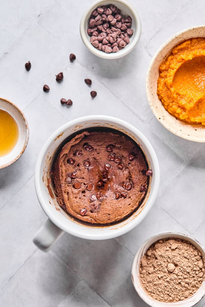 Overhead shot of a chocolate mug cake with chocolate chips. Around the mug is a bowl of pumpkin puree, a bowl of chocolate protein powder, a bowl of maple syrup, and a bowl of mini chocolate chips with some around the bowl.