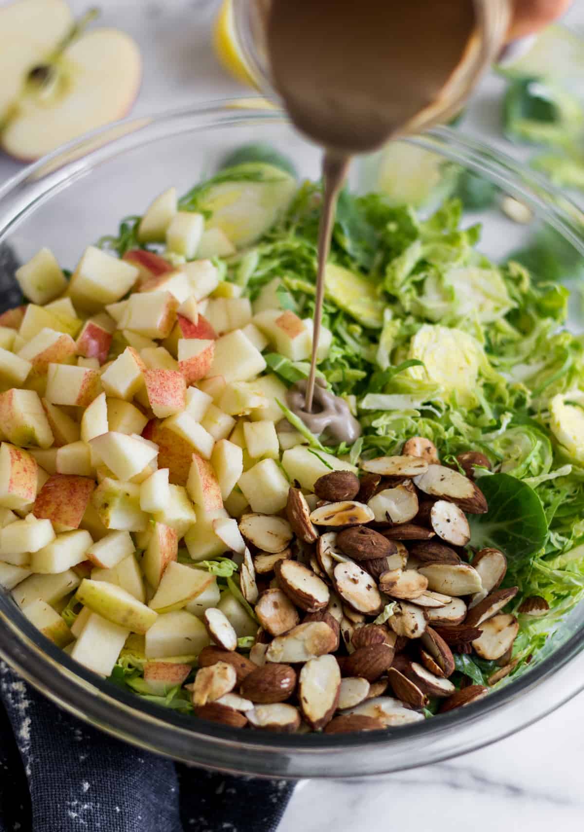 Glass bowl with shredded brussels sprouts, chopped apple, and toasted almonds and creamy balsamic dressing being poured into it